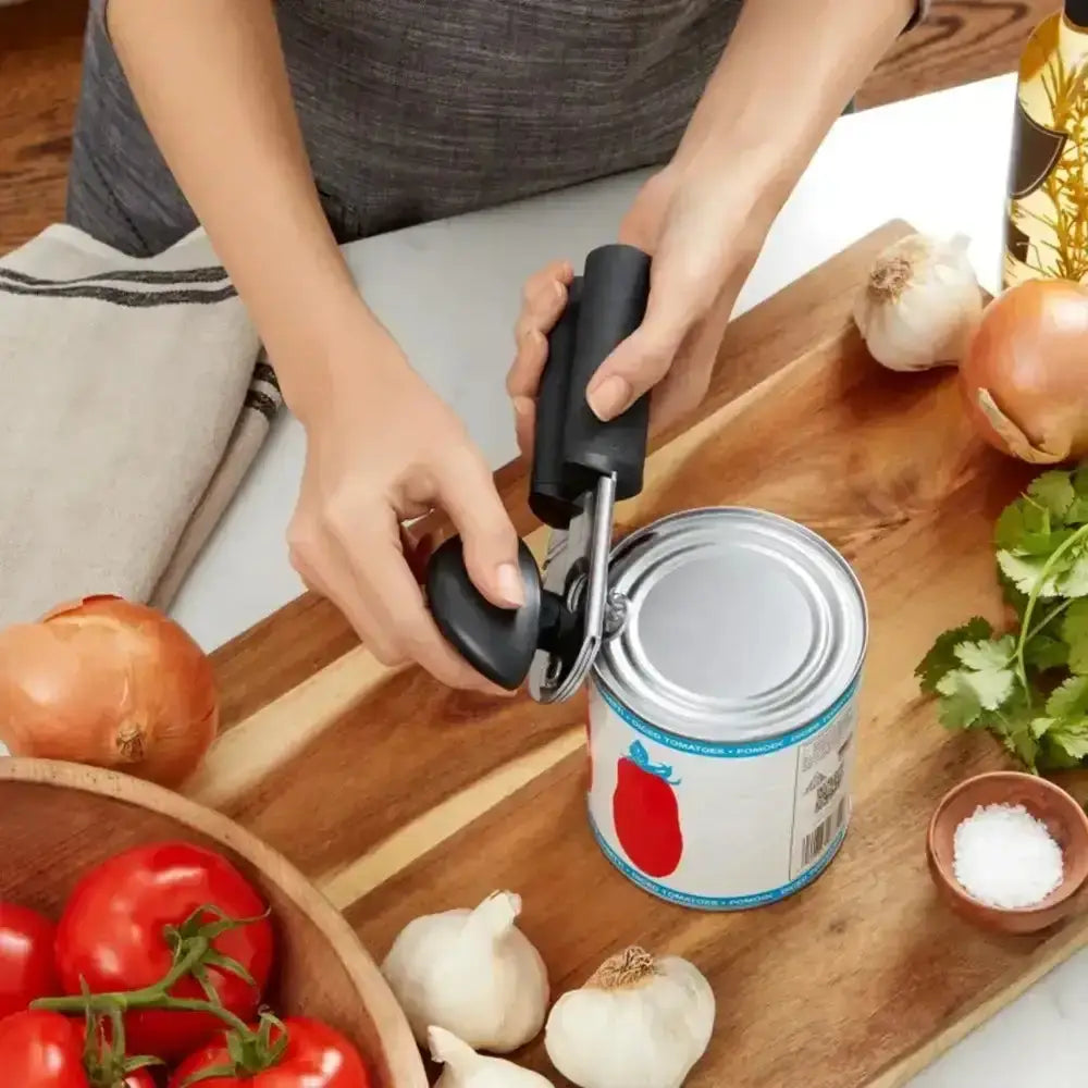 Person using a can opener on a can with vegetables and kitchen items on a wooden board.