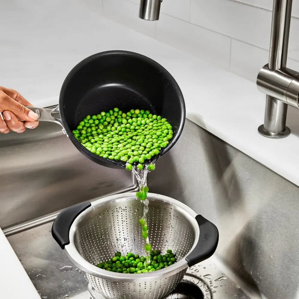Green peas being poured from a black pot into a silver colander in a kitchen sink.