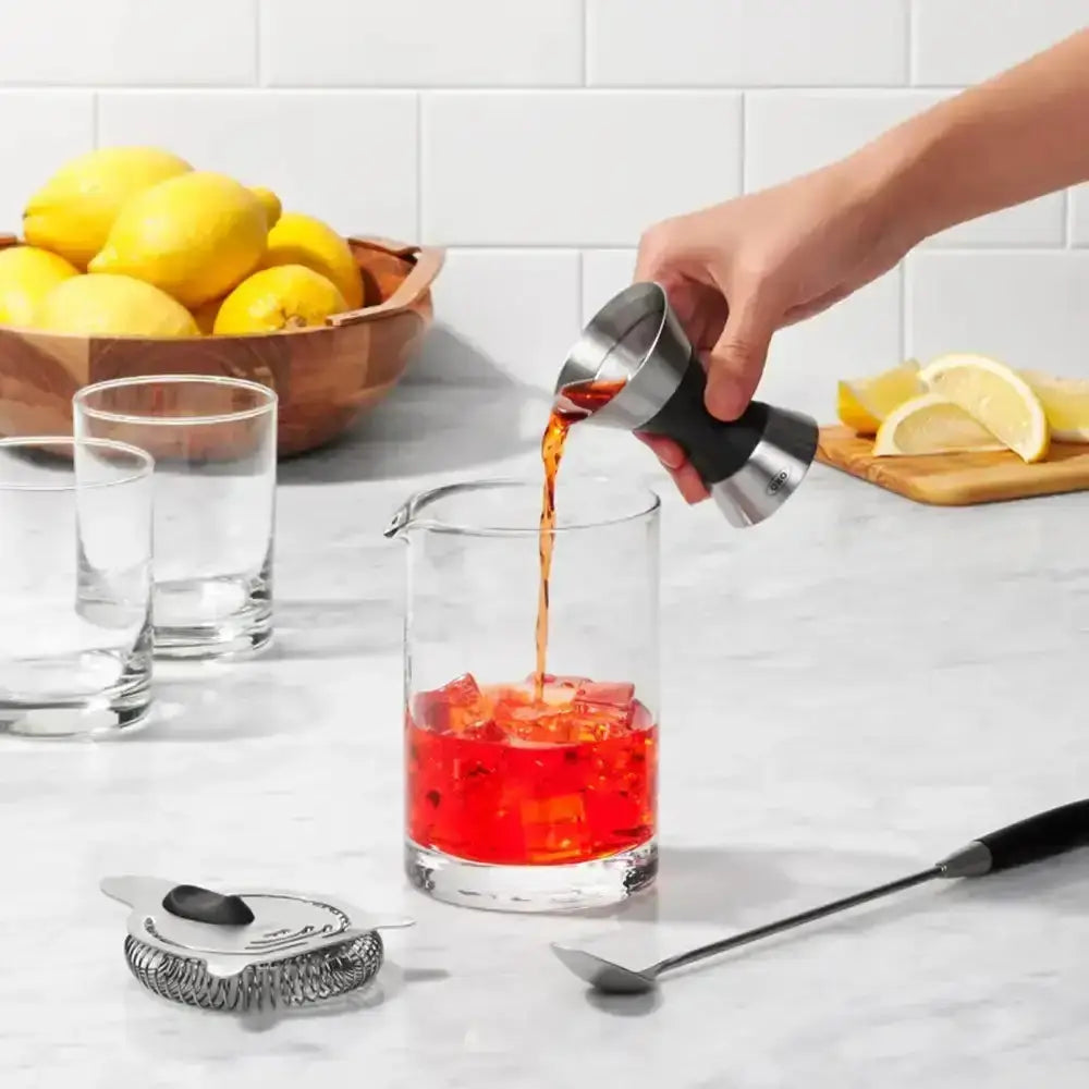 Person pouring a red liquid from a measuring cup into a glass on a kitchen counter with lemons and glasses in the background.