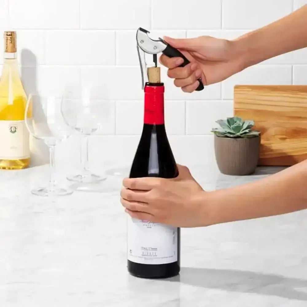Person opening a bottle of red wine with a corkscrew in a kitchen setting.