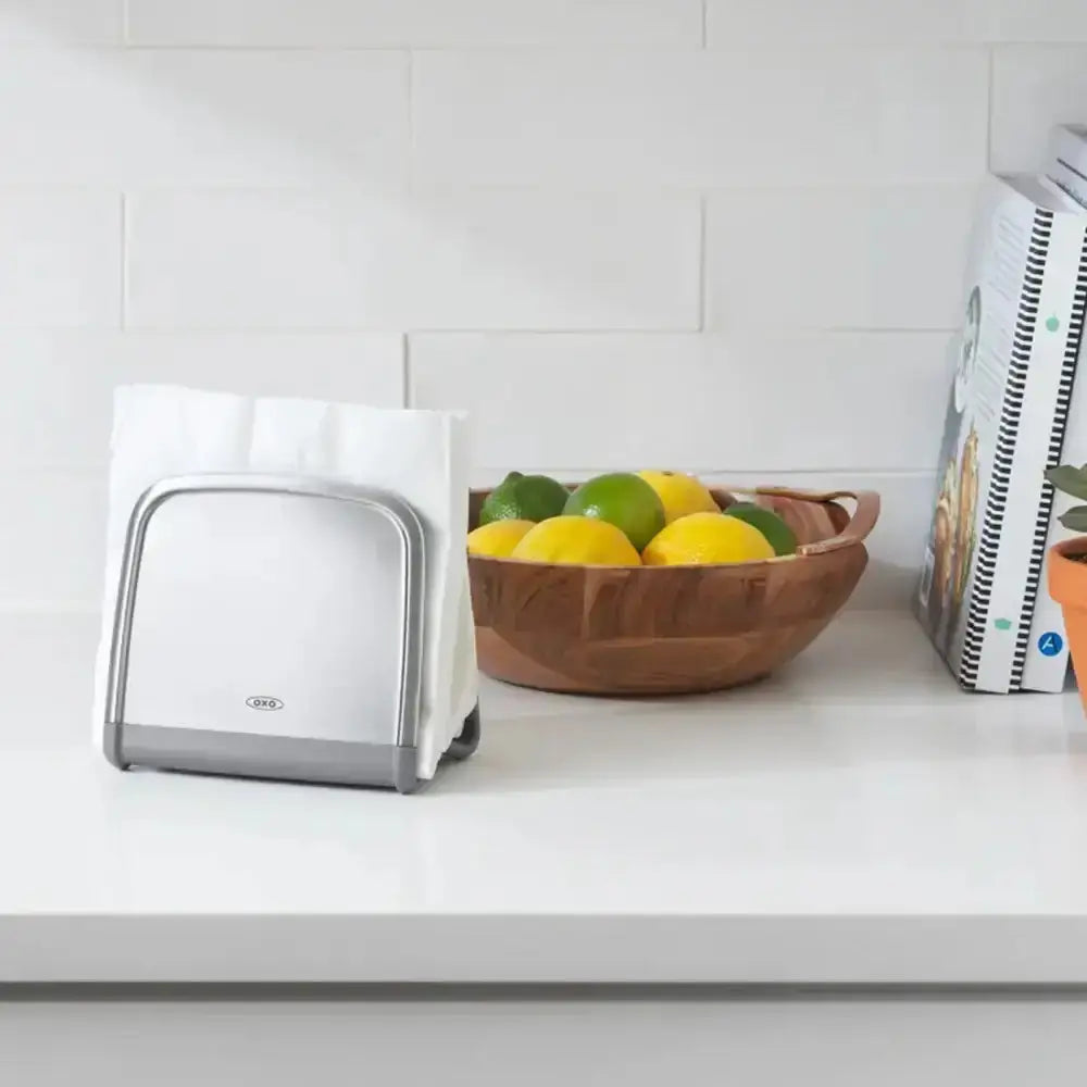 White paper towel holder on a kitchen counter with a bowl of fruit and books in the background.