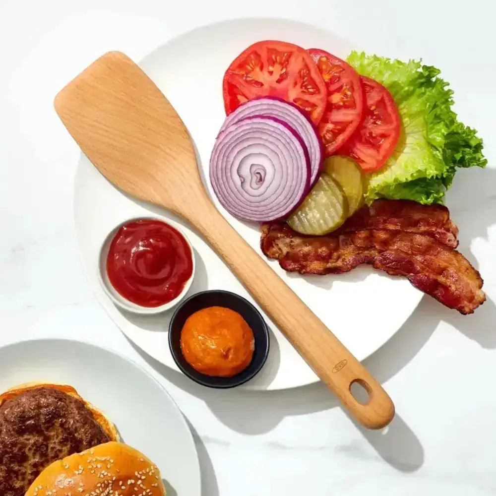 Burger ingredients including lettuce, tomato, onion, pickle, and bacon on a white plate with a wooden spatula and condiments.