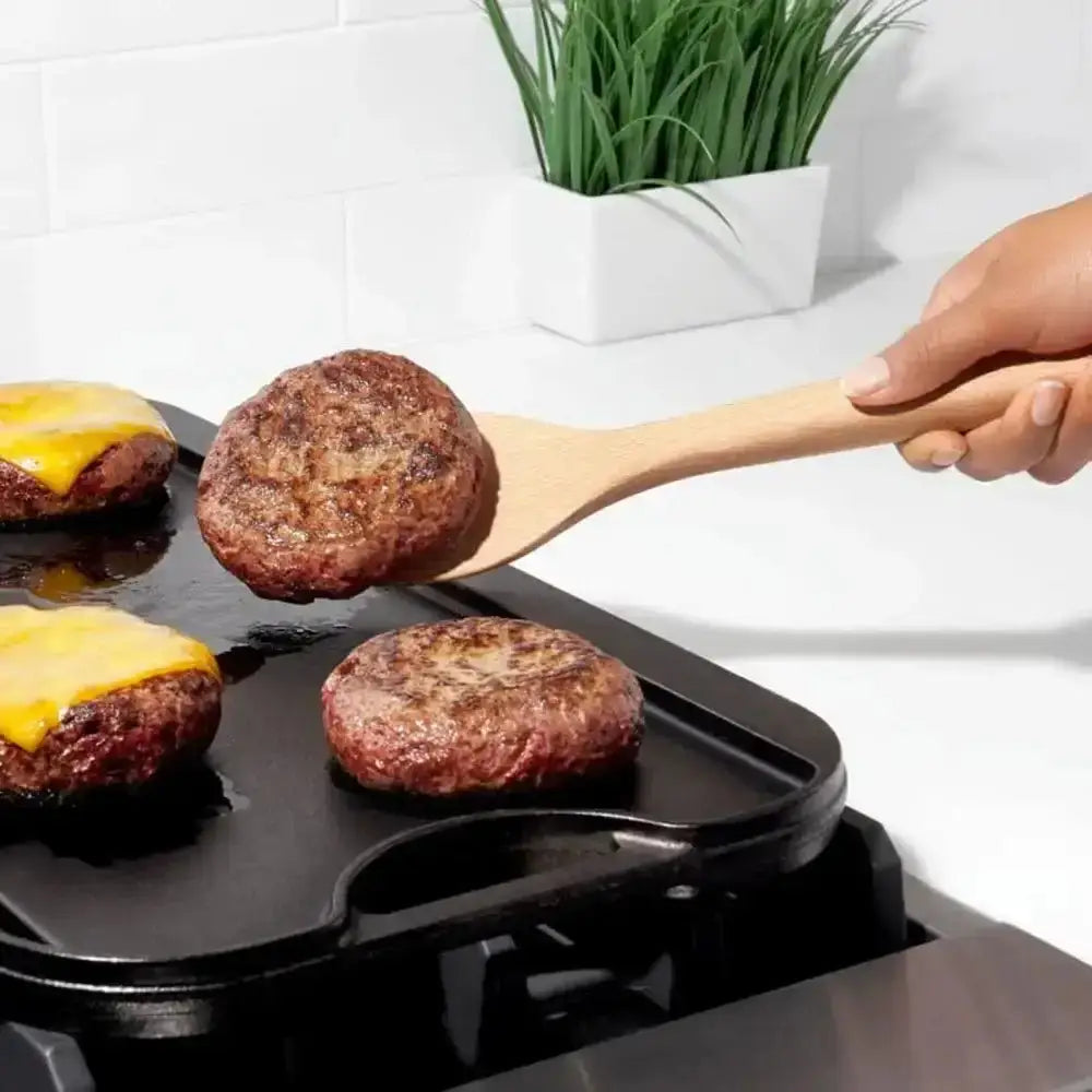 Hamburgers being cooked on a grill with a wooden spatula, plant in the background.