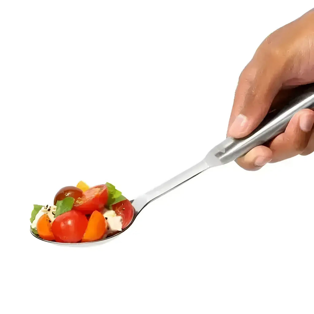 A hand holds an oxo stainless steel serving spoon filled with cherry tomatoes basil and crumbled cheese