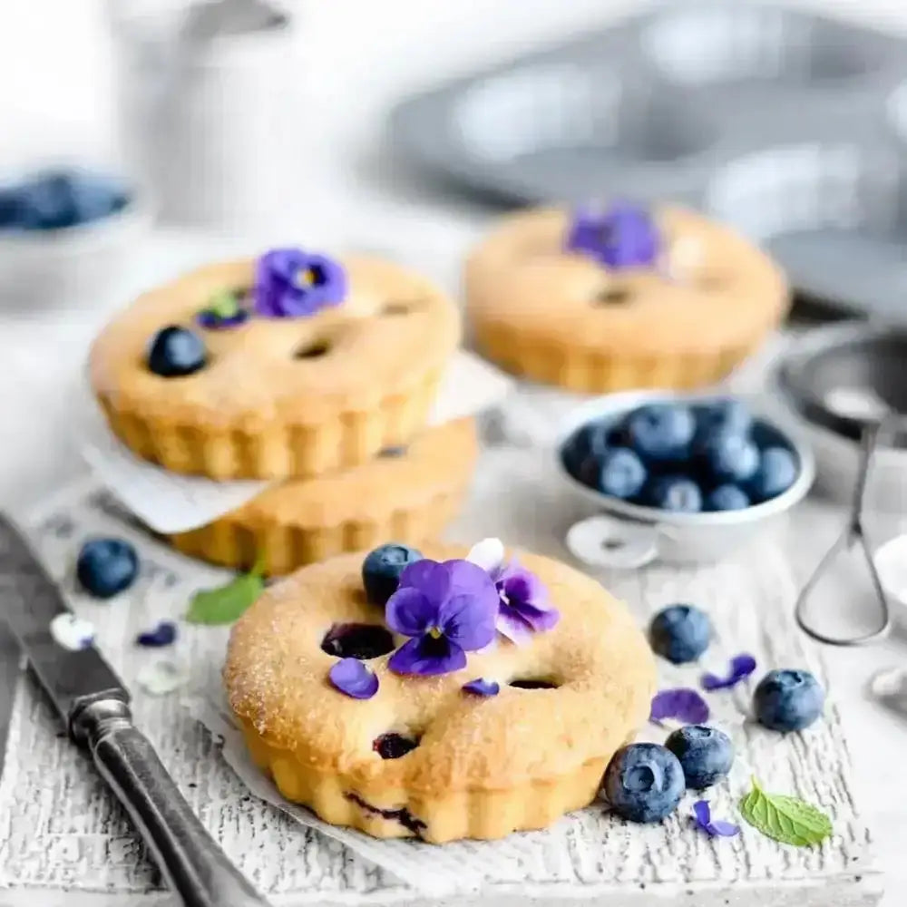 Blueberry muffins with purple flowers on a white surface with blueberries and baking tools in the background.