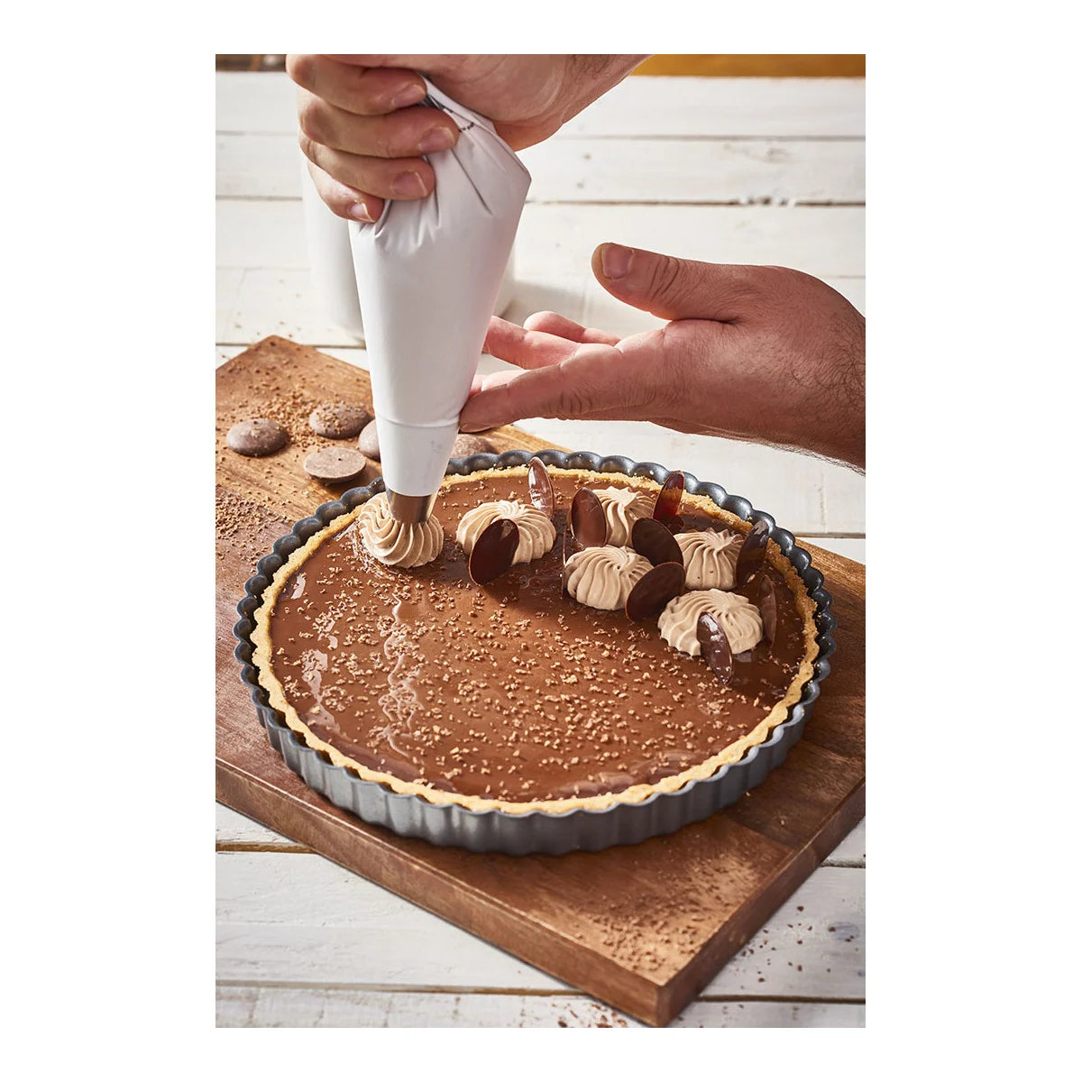 Person decorating a chocolate tart with a piping bag on a wooden board.