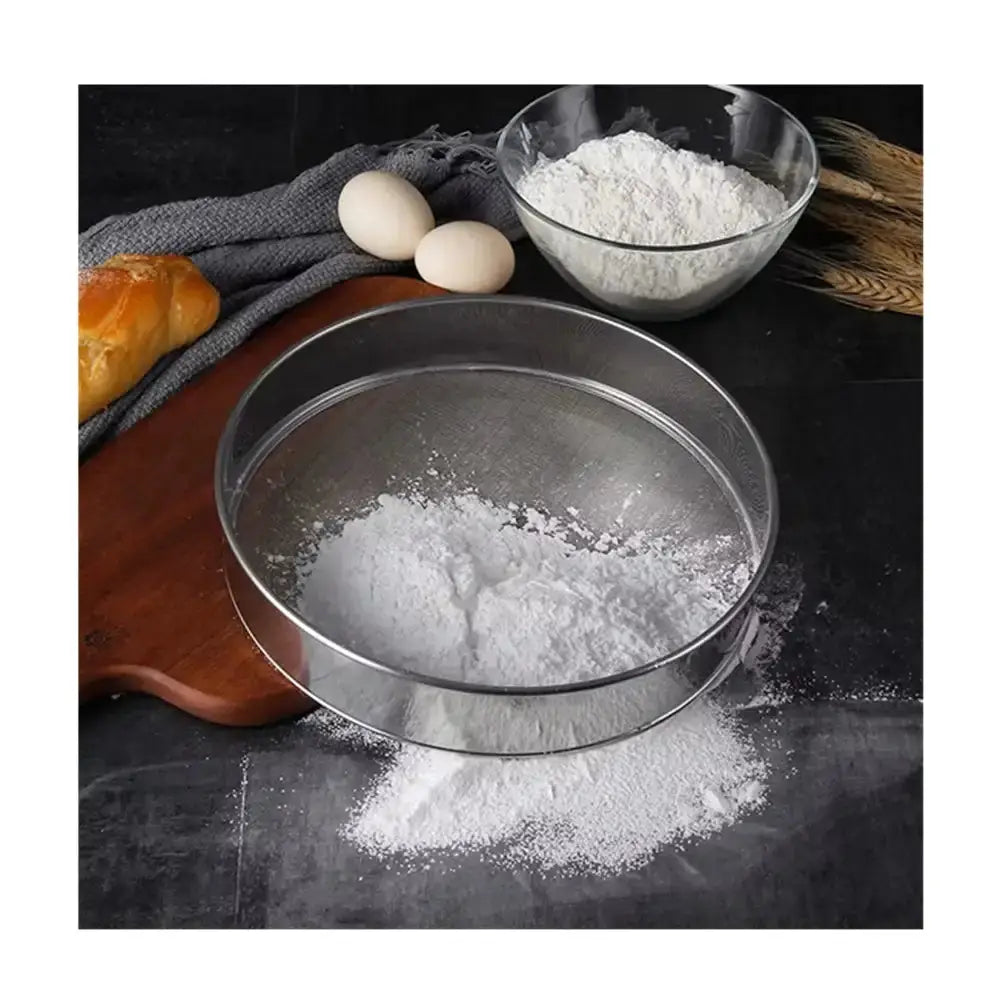 Metal sieve with flour on a dark surface with eggs and bread in the background