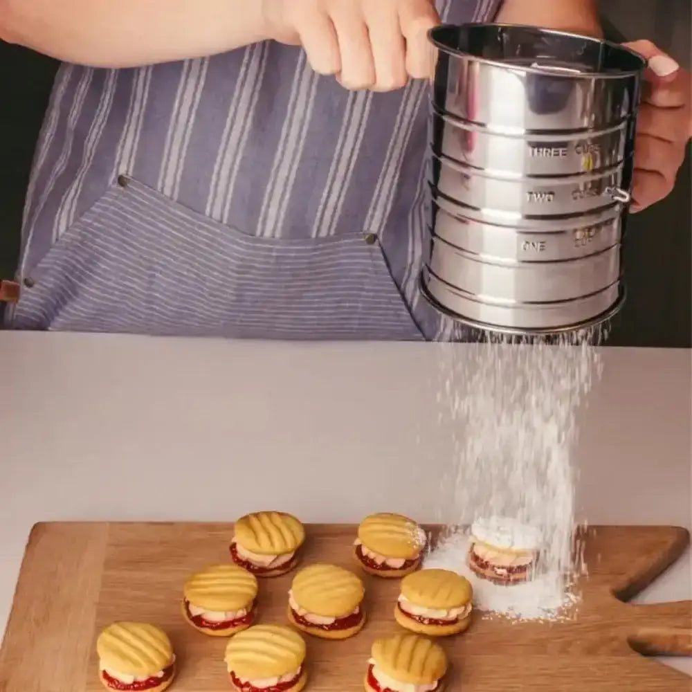 Person sifting powdered sugar over small sandwiches on a wooden board.
