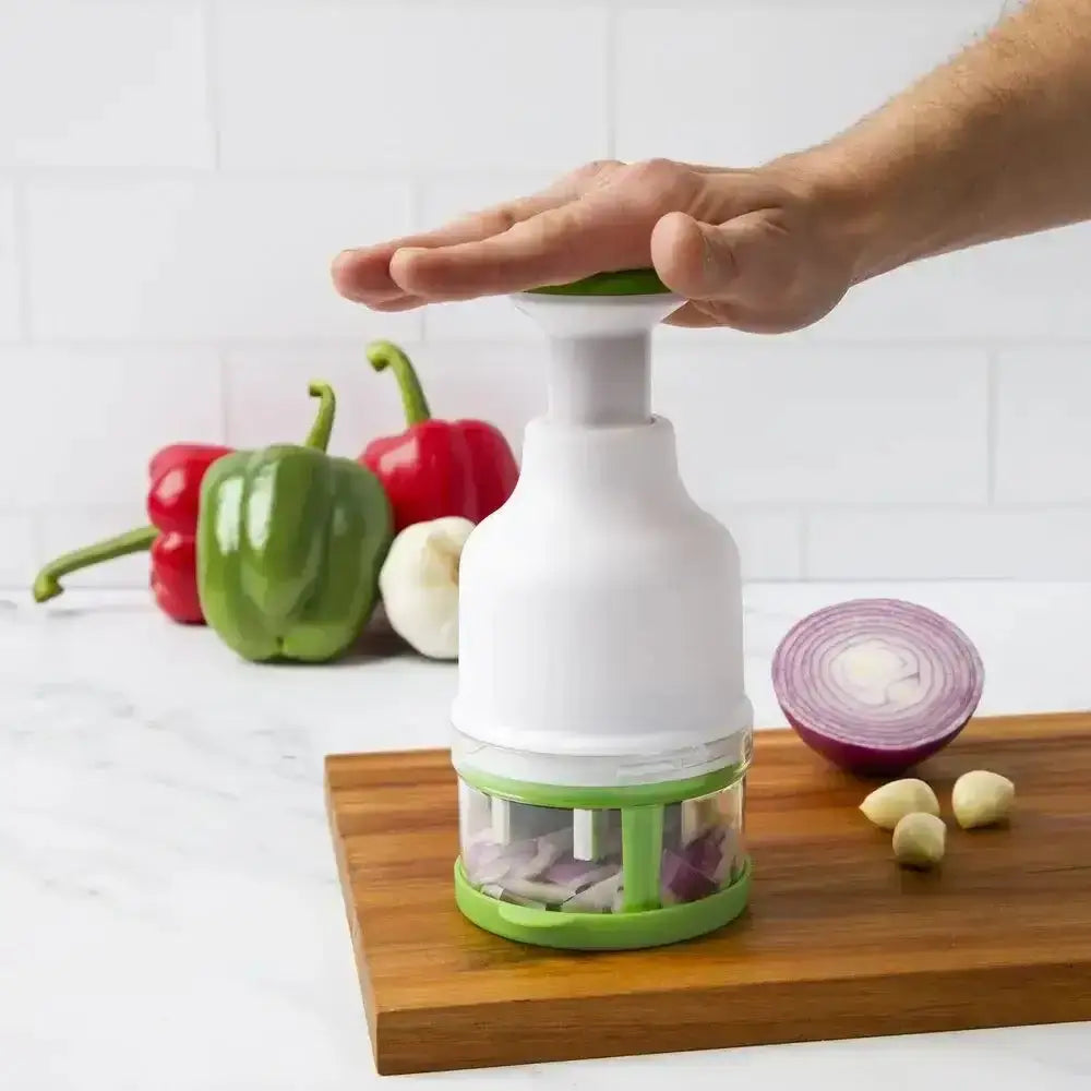 Hand using a white and green kitchen tool on a wooden cutting board with vegetables in the background.