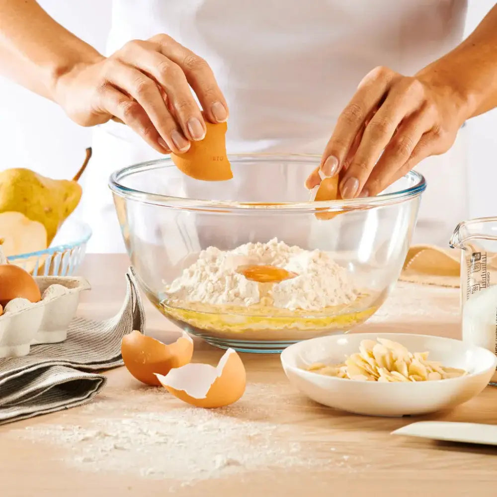 Person cracking eggs into a bowl of flour on a wooden table with kitchen items around.