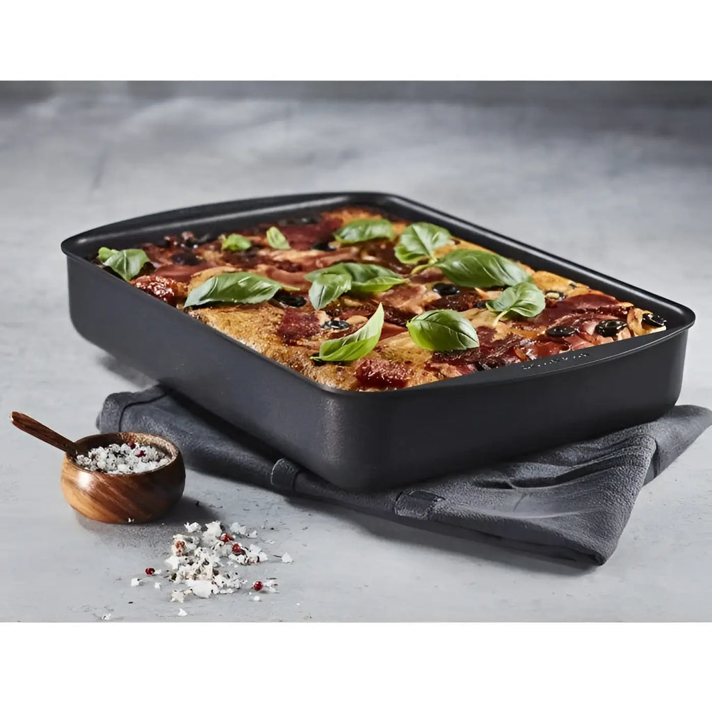 Baked dish in a black rectangular baking tray on a gray surface with a small wooden bowl of herbs.