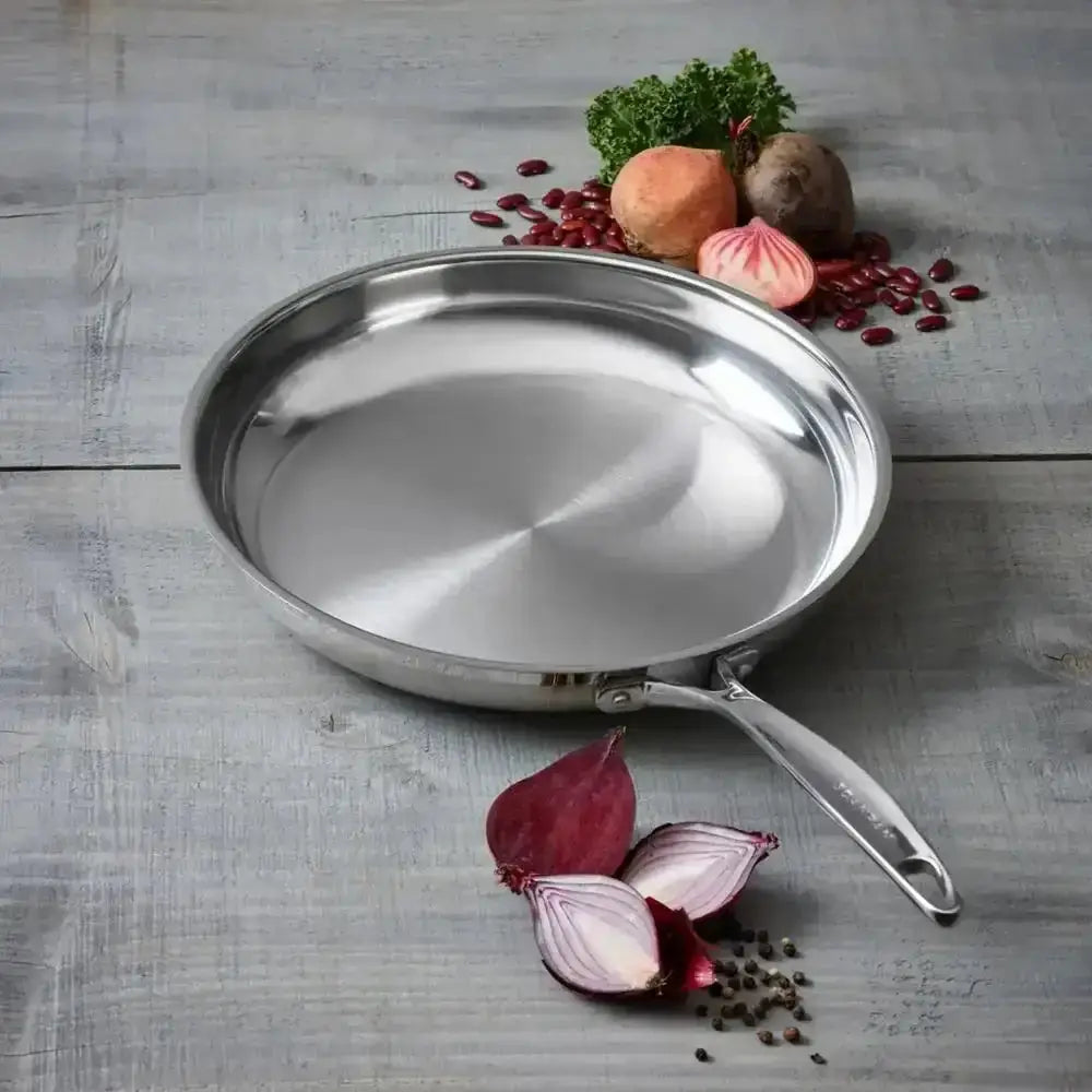 Stainless steel frying pan on a wooden surface with vegetables and spices.