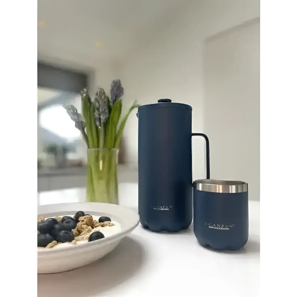 Blue ceramic pitcher and cup on a kitchen counter with a bowl of cereal and flowers in the background.