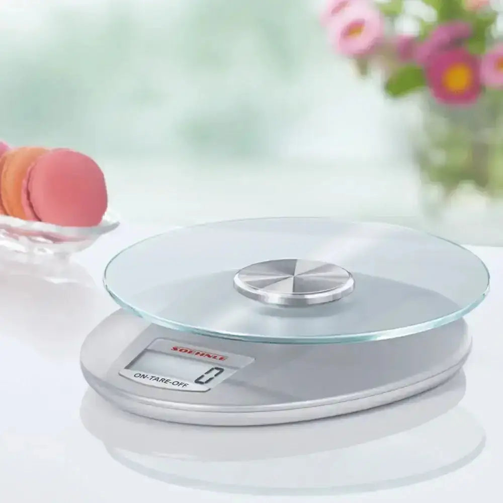 Digital kitchen scale on a white surface with blurred flowers and macarons in the background