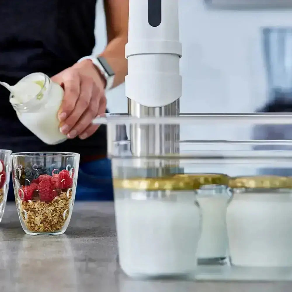 Person pouring yogurt into a glass with granola and berries, using a kitchen appliance.