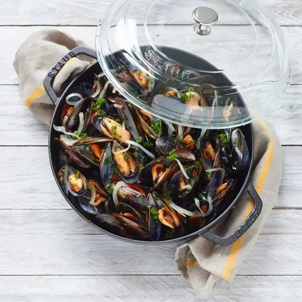 Black enameled cast iron staub cocotte with glass lid filled with cooked mussels and parsley for stovetop cooking
