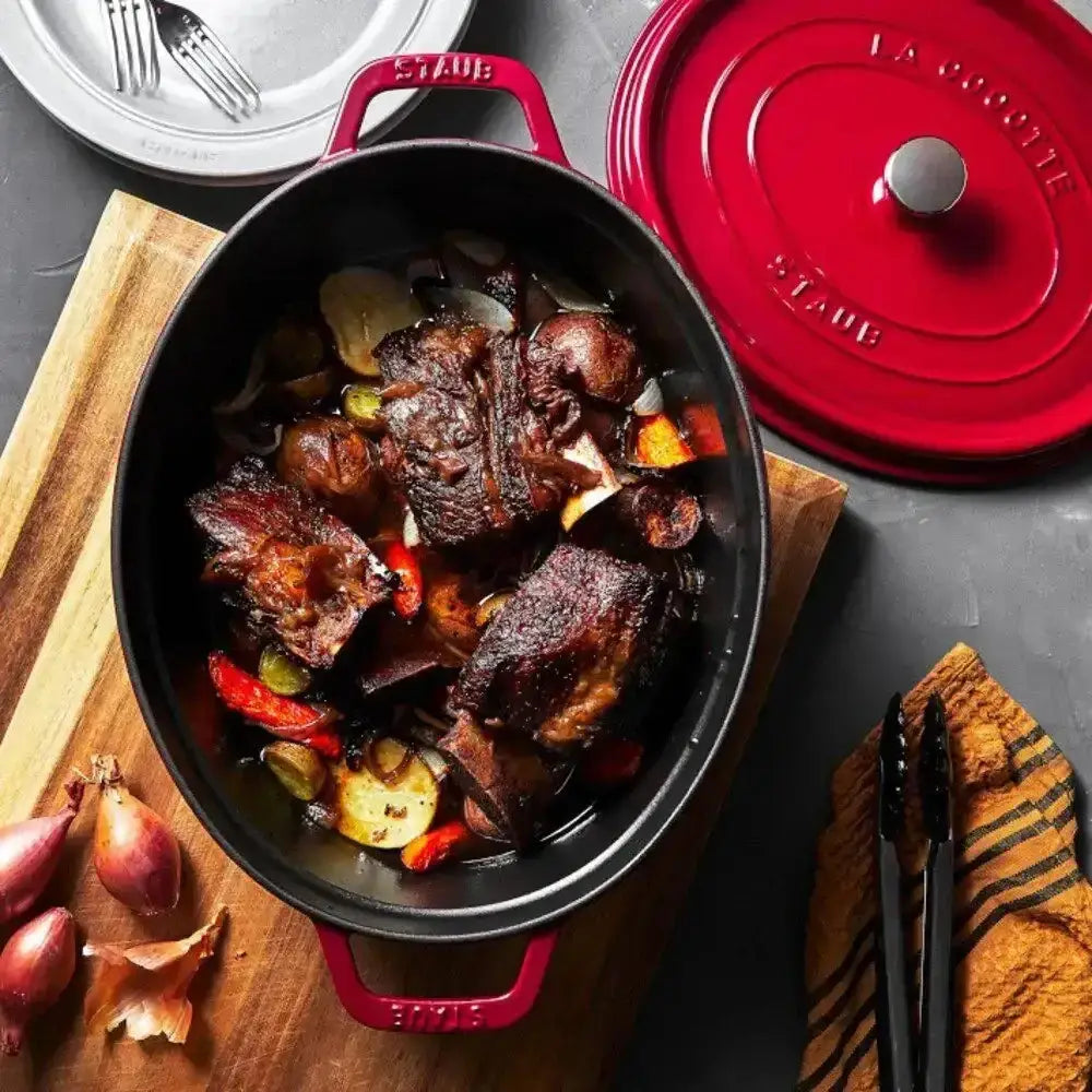 Red Staub pot with braised meat and vegetables on a wooden board, surrounded by kitchen items.