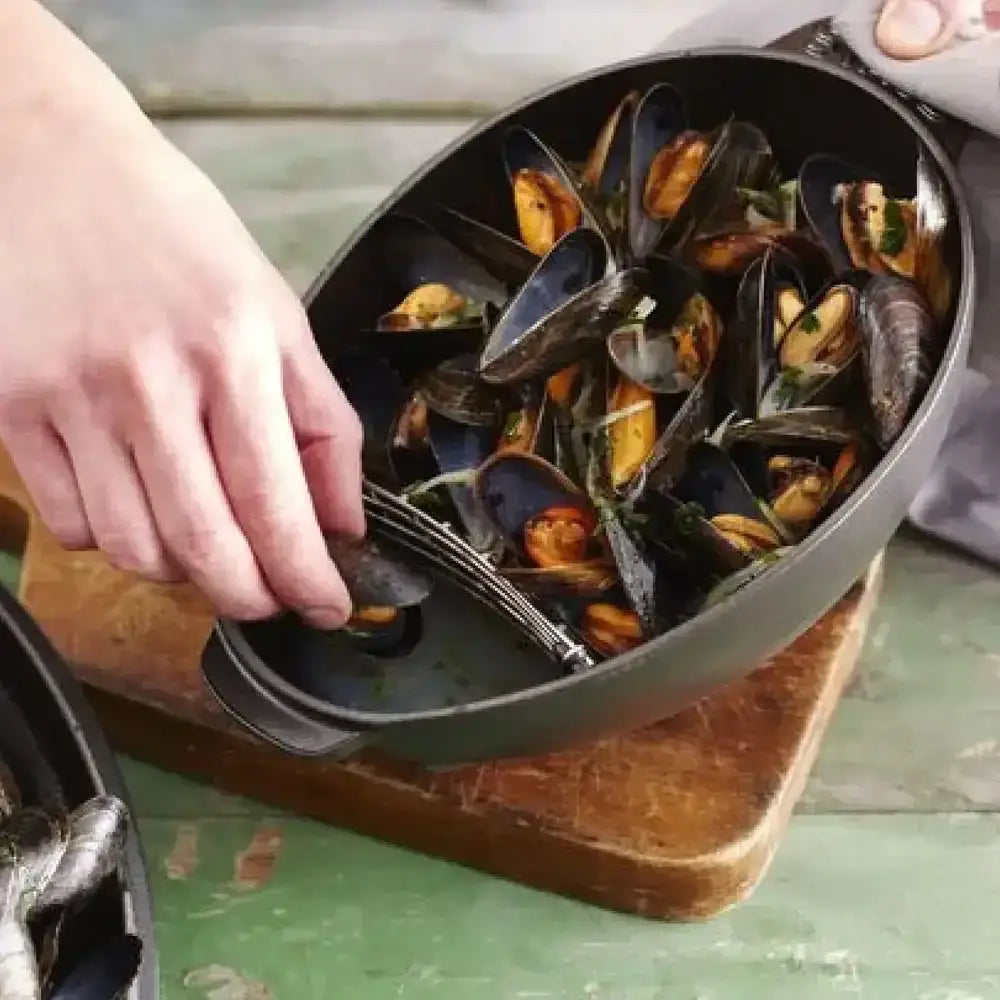 Staub oval cast iron mussel pot 2l black ceramic baking dish filled with cooked mussels and green herb garnish