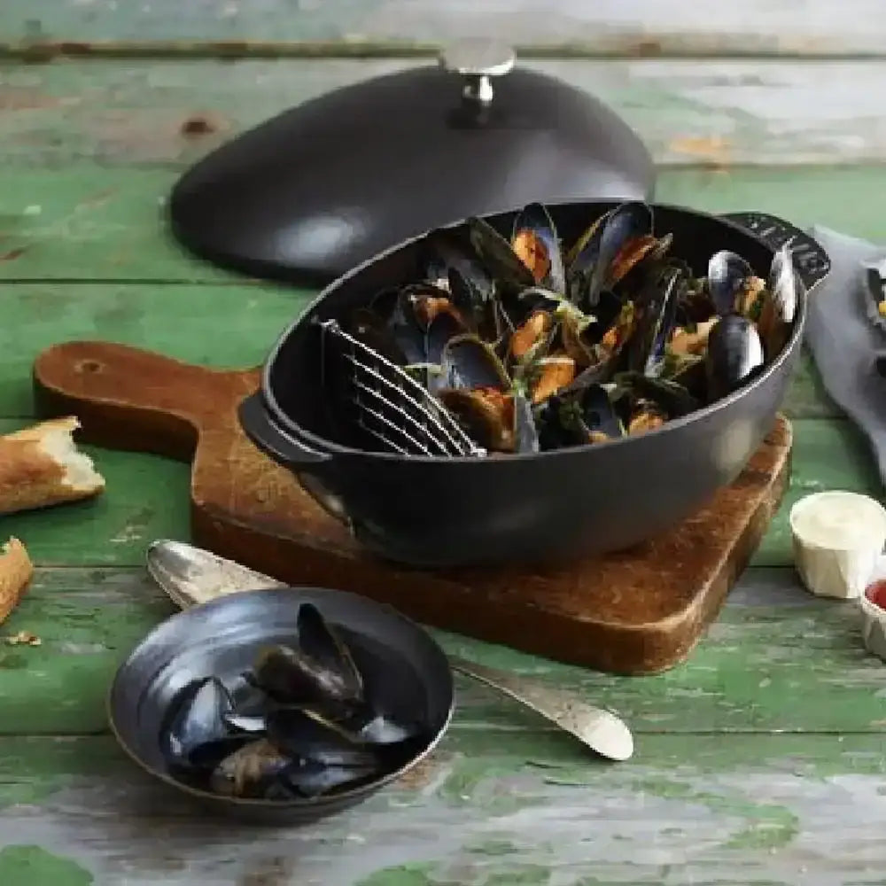 Black cast iron staub oval dutch oven with lid and handles filled with cooked mussels 2l capacity