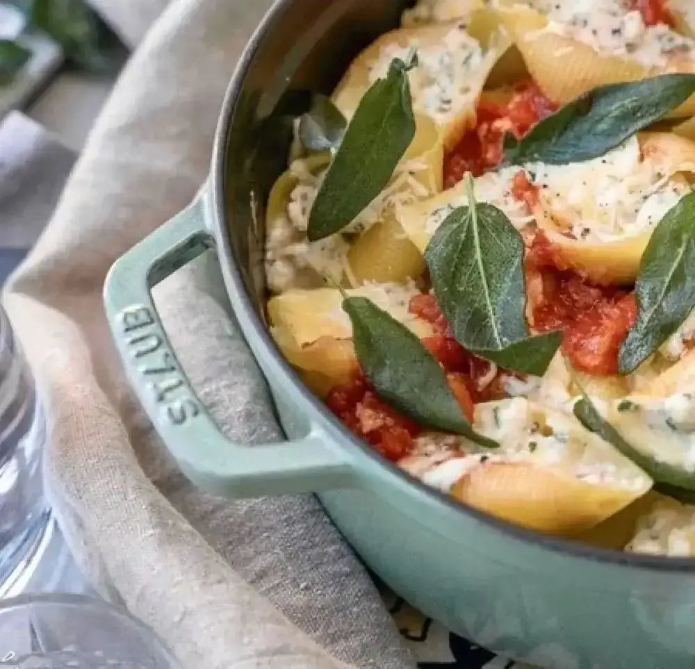 Staub sage green enameled cast iron dutch oven filled with stuffed pasta shells topped with tomato sauce and basil
