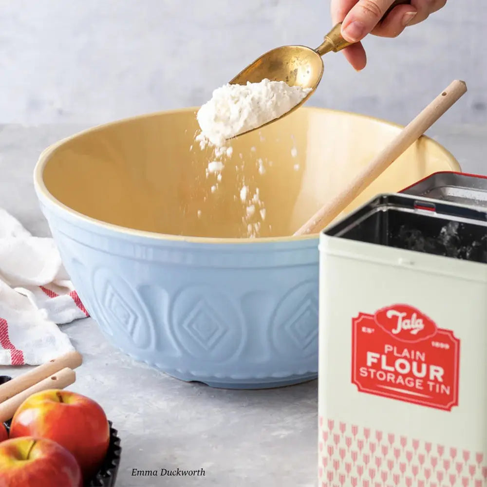 Person adding flour to a blue mixing bowl with a Tala flour storage tin in the background.