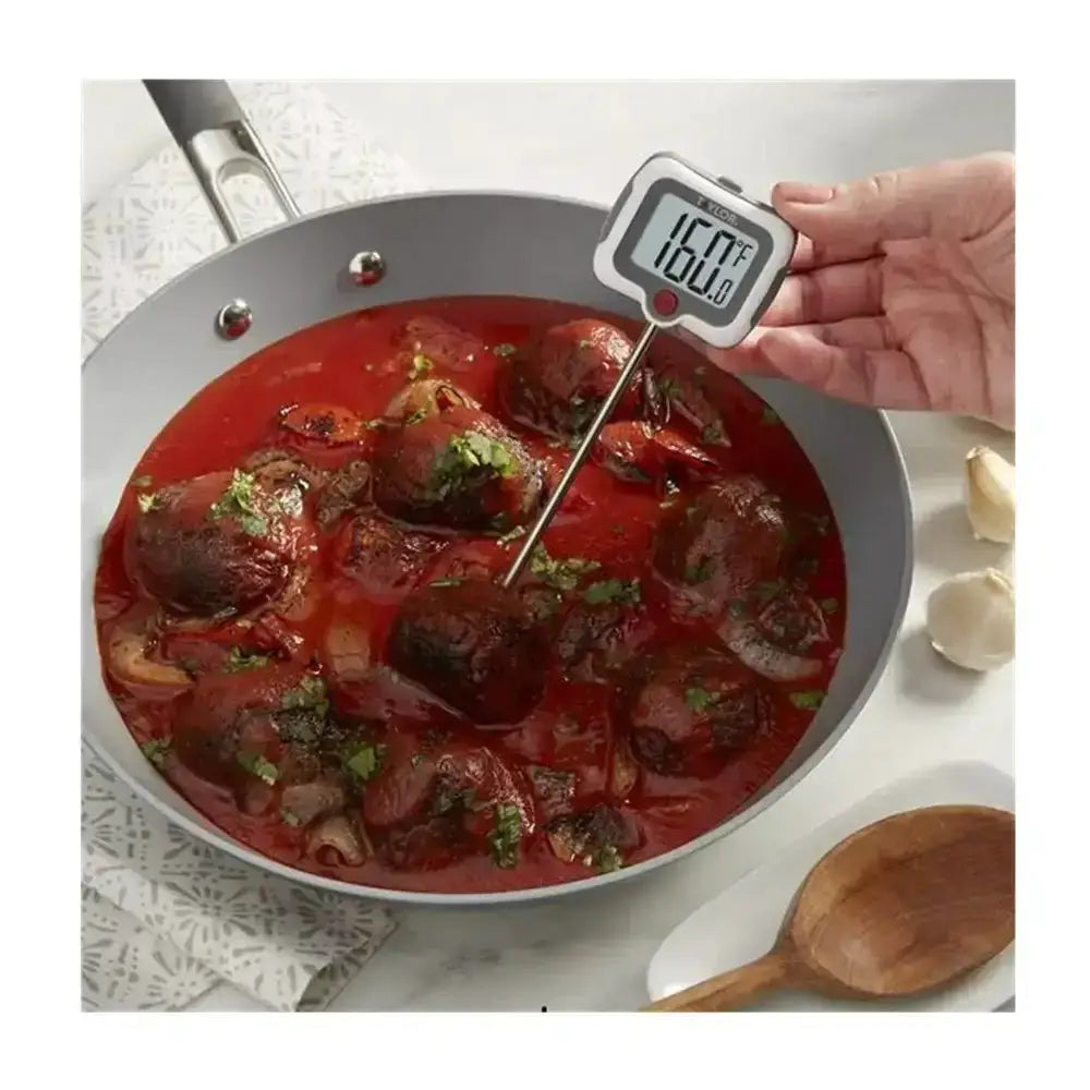 Digital thermometer inserted into a dish of food with a wooden spoon and garlic cloves on a white surface.