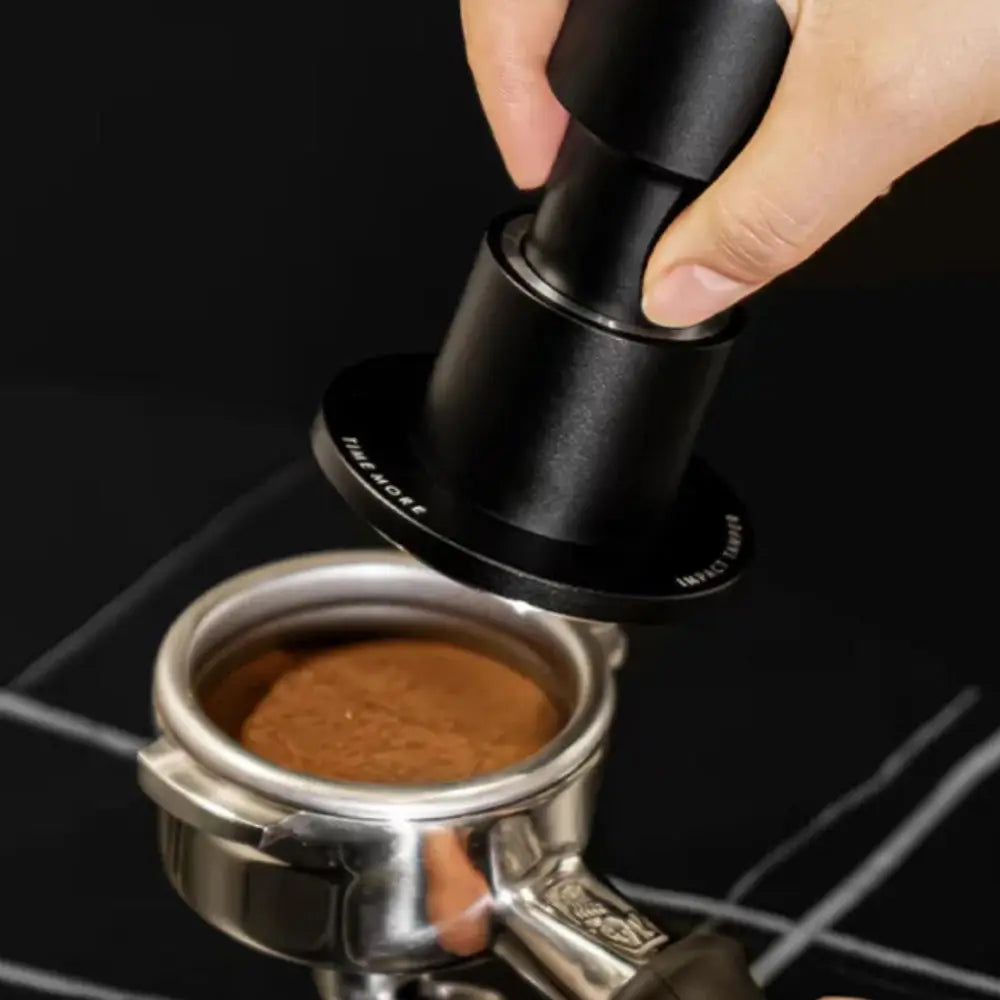 Hand using a black tamper to compress coffee grounds in a silver espresso portafilter against a black background.