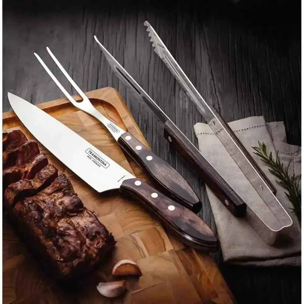 Set of knives and carving tools on a wooden cutting board with cooked meat and garlic.
