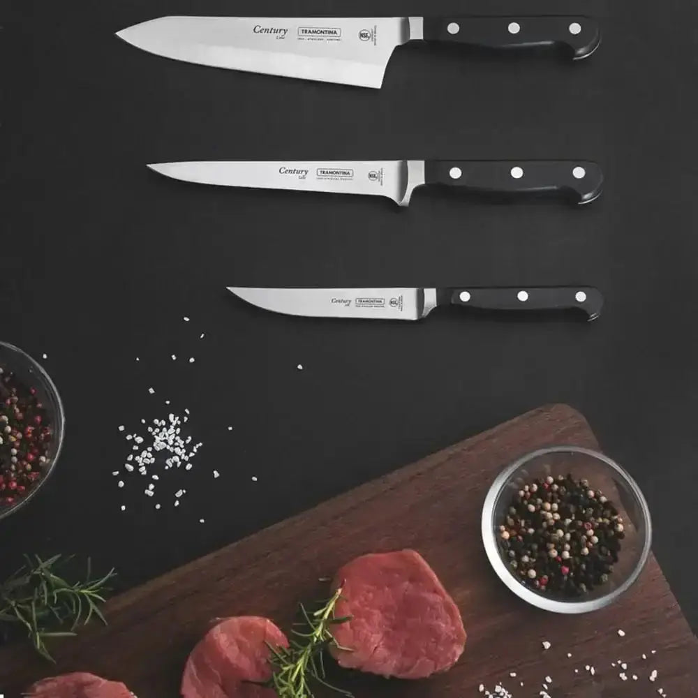 Three knives on a dark surface with a wooden cutting board and ingredients below.