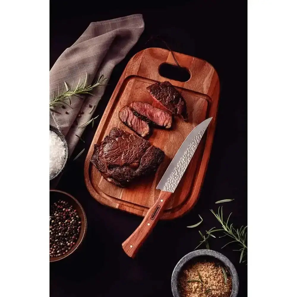 Wooden cutting board with cooked meat, a knife, and seasonings on a dark surface.