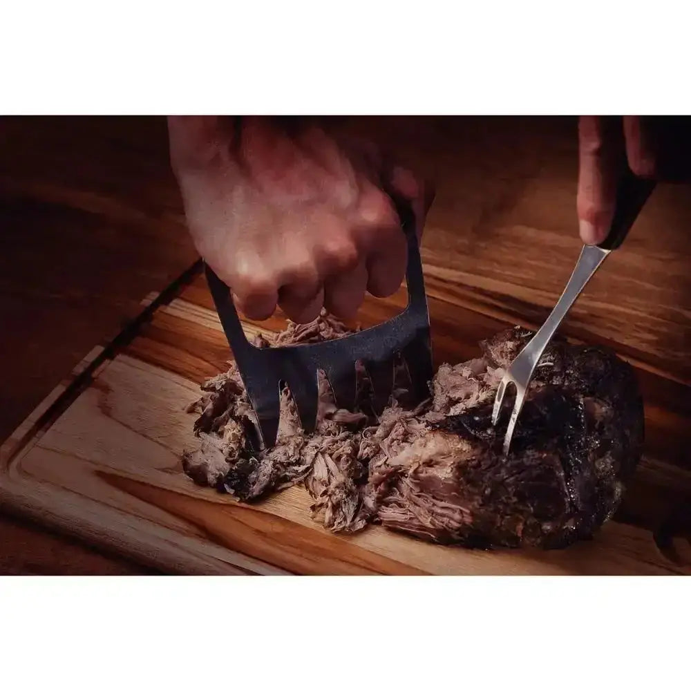 Stainless steel meat claw and fork shredding cooked meat on wooden cutting board