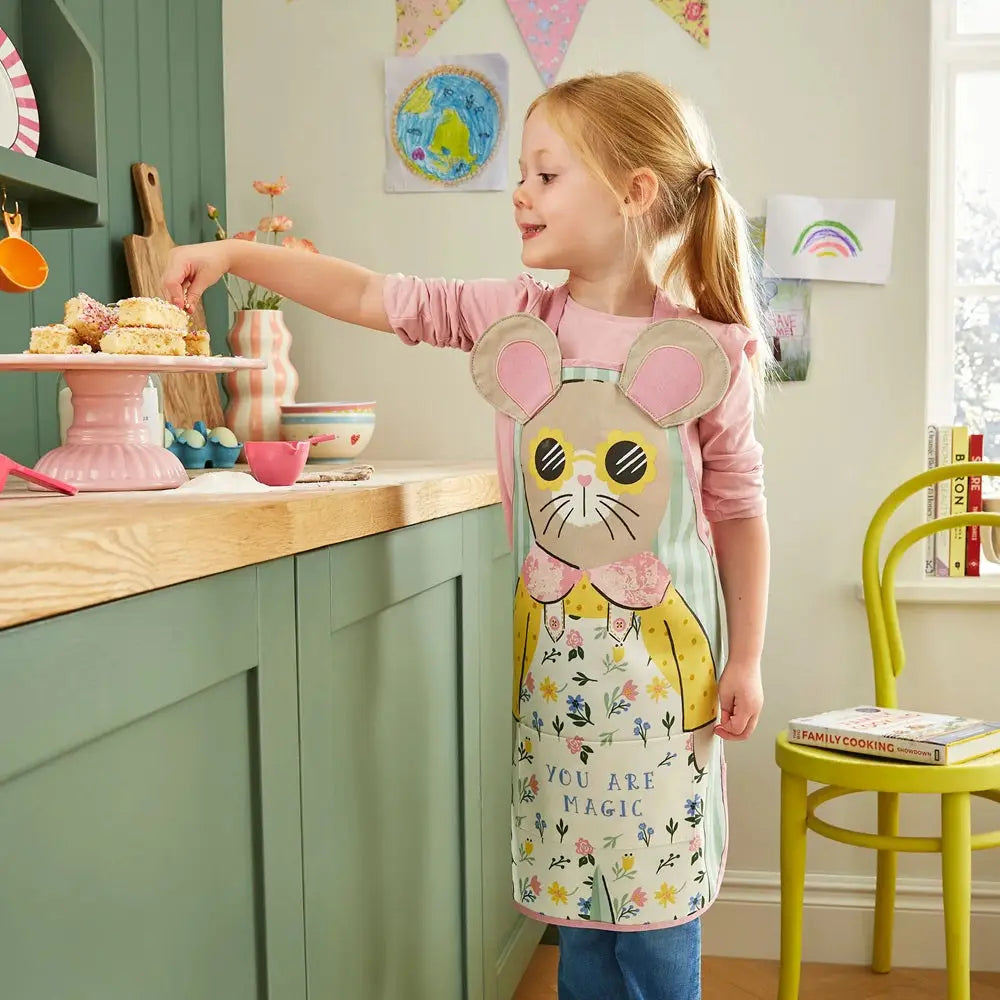Child in a kitchen wearing a mouse-themed apron, reaching for a cake.