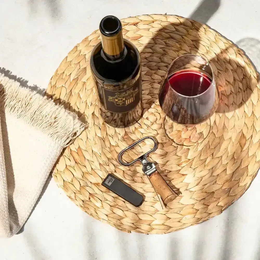 Wine bottle, glass with red wine, and opener on a woven mat