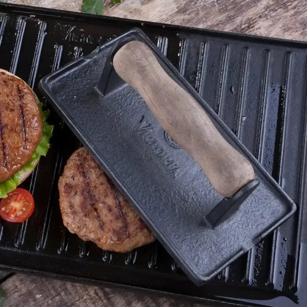 Black metal burger press on a grill with two burgers and a tomato