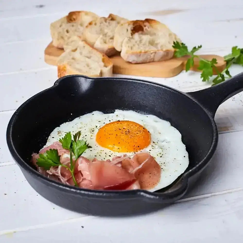 Fried egg with ham in a black skillet on a wooden surface with bread in the background