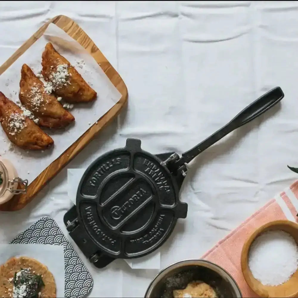 Cast iron tortilla press on a white surface with fried food items and a wooden tray.