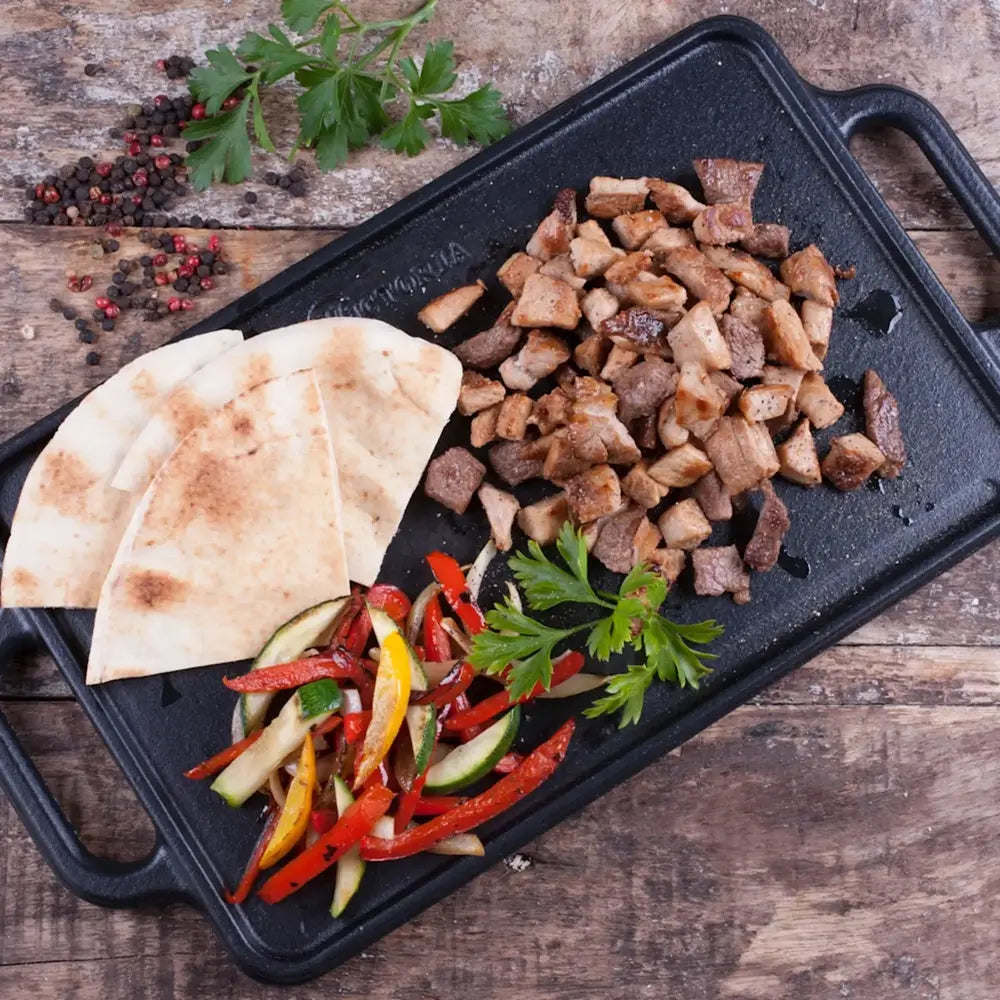 Black grill pan with cooked meat, vegetables, and flatbread on a wooden surface.