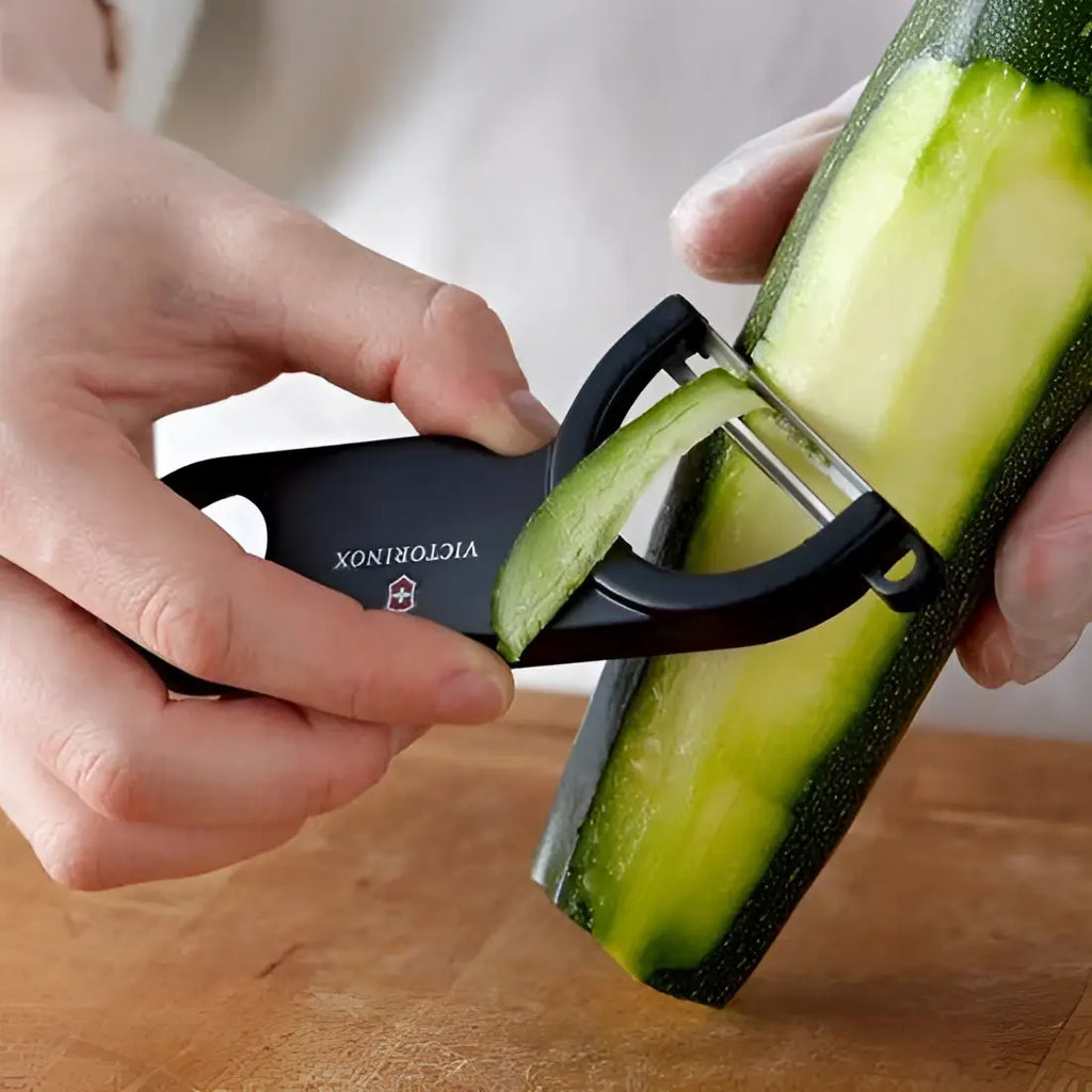 Person using a Victorinox vegetable peeler to slice a zucchini on a wooden surface.