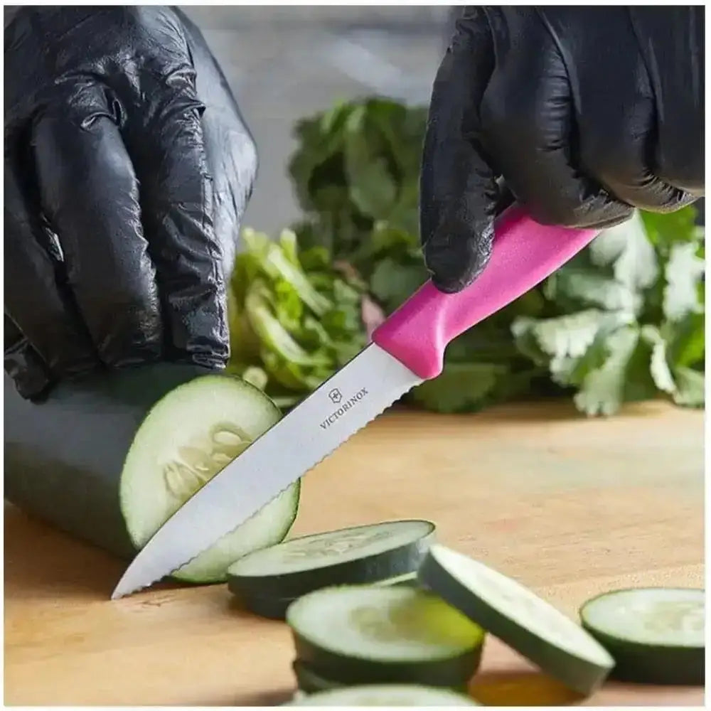 Person wearing black gloves using a pink Victorinox knife to slice cucumbers on a wooden cutting board.