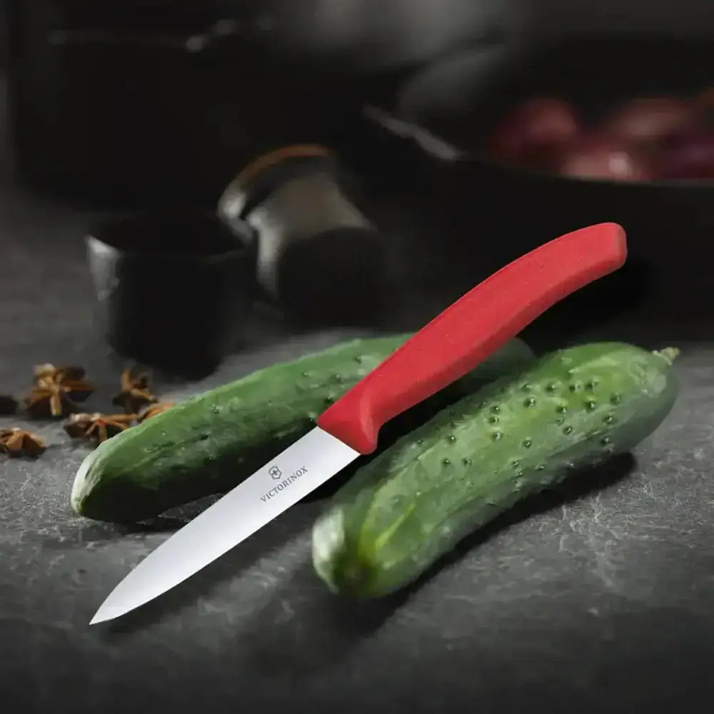 Paring knife with red handle on a cucumber on a dark surface