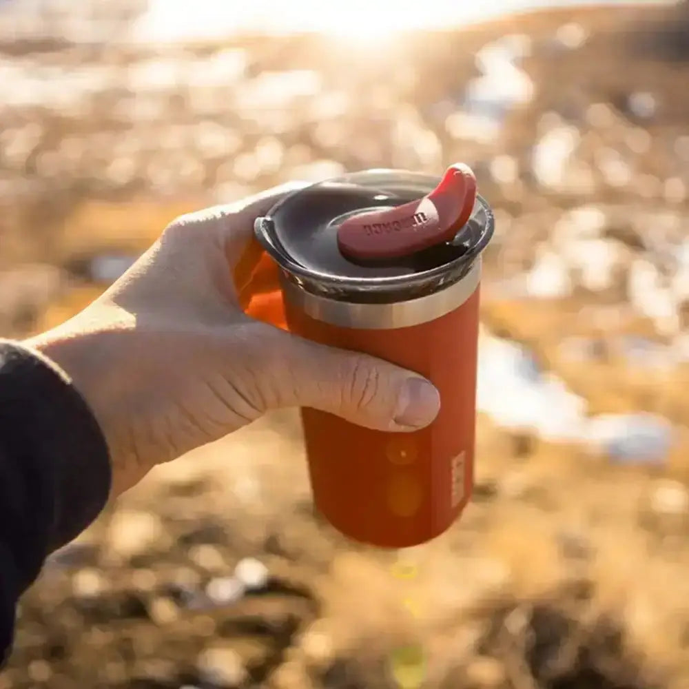 Hand holding an orange insulated mug with a red handle against a blurred natural background