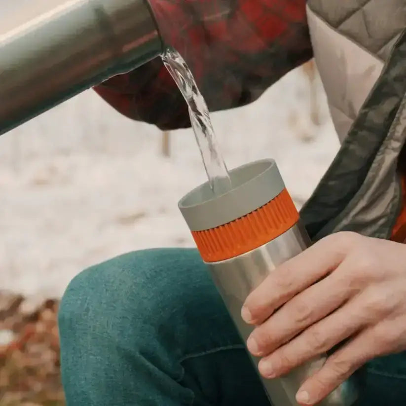 Person pouring water from a metal container into a silver and orange insulated cup outdoors.