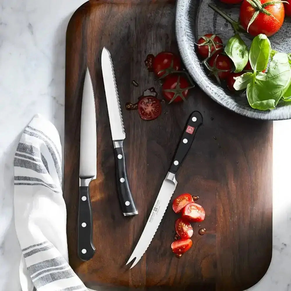 Three knives on a wooden cutting board with sliced tomatoes and basil.