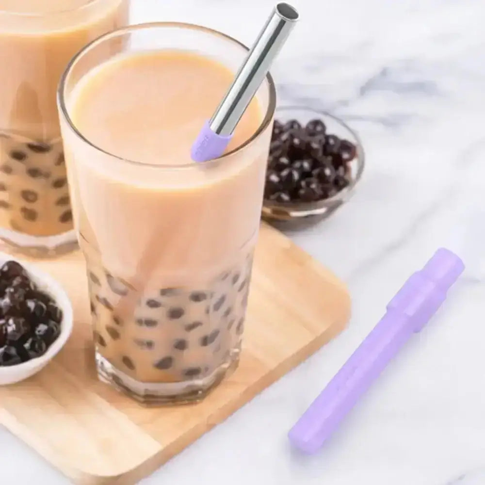 Glass of bubble tea with a purple straw on a wooden coaster, surrounded by bowls of boba and another glass of bubble tea.
