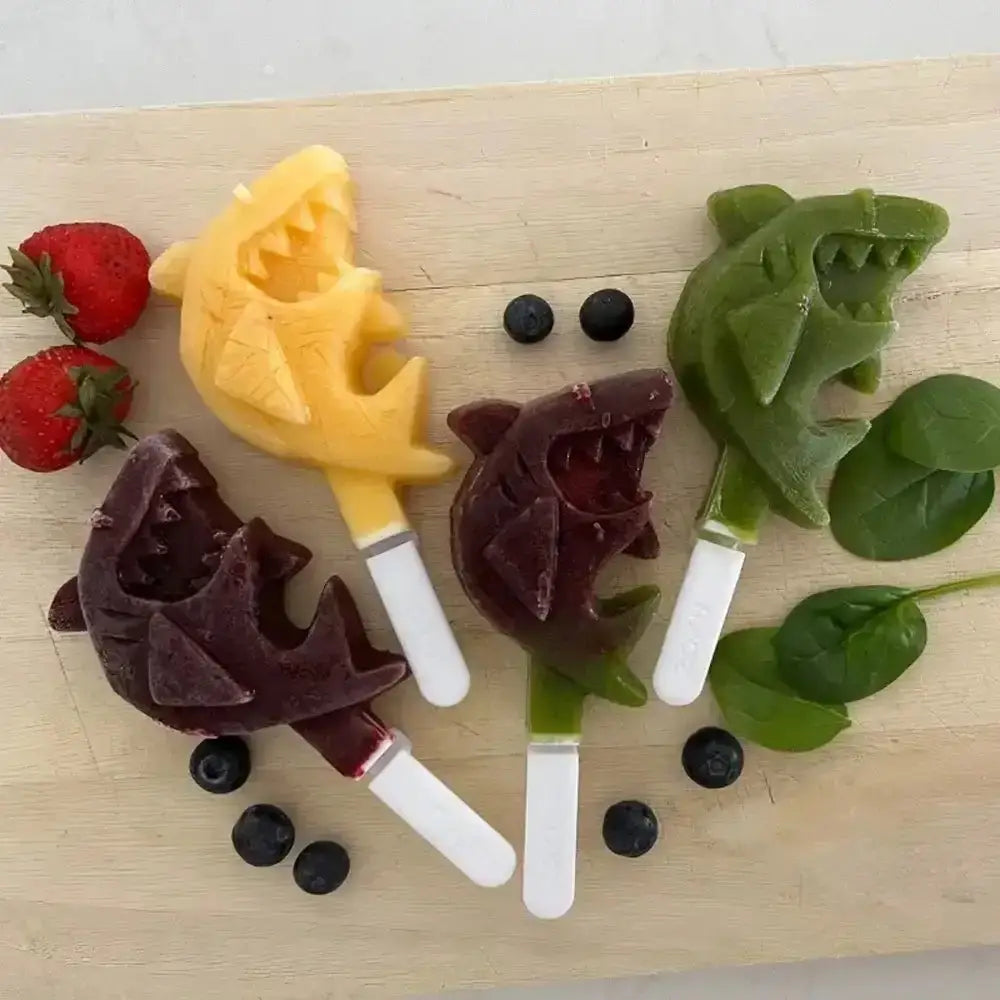 Four popsicles shaped like animals on a wooden board with berries and spinach.
