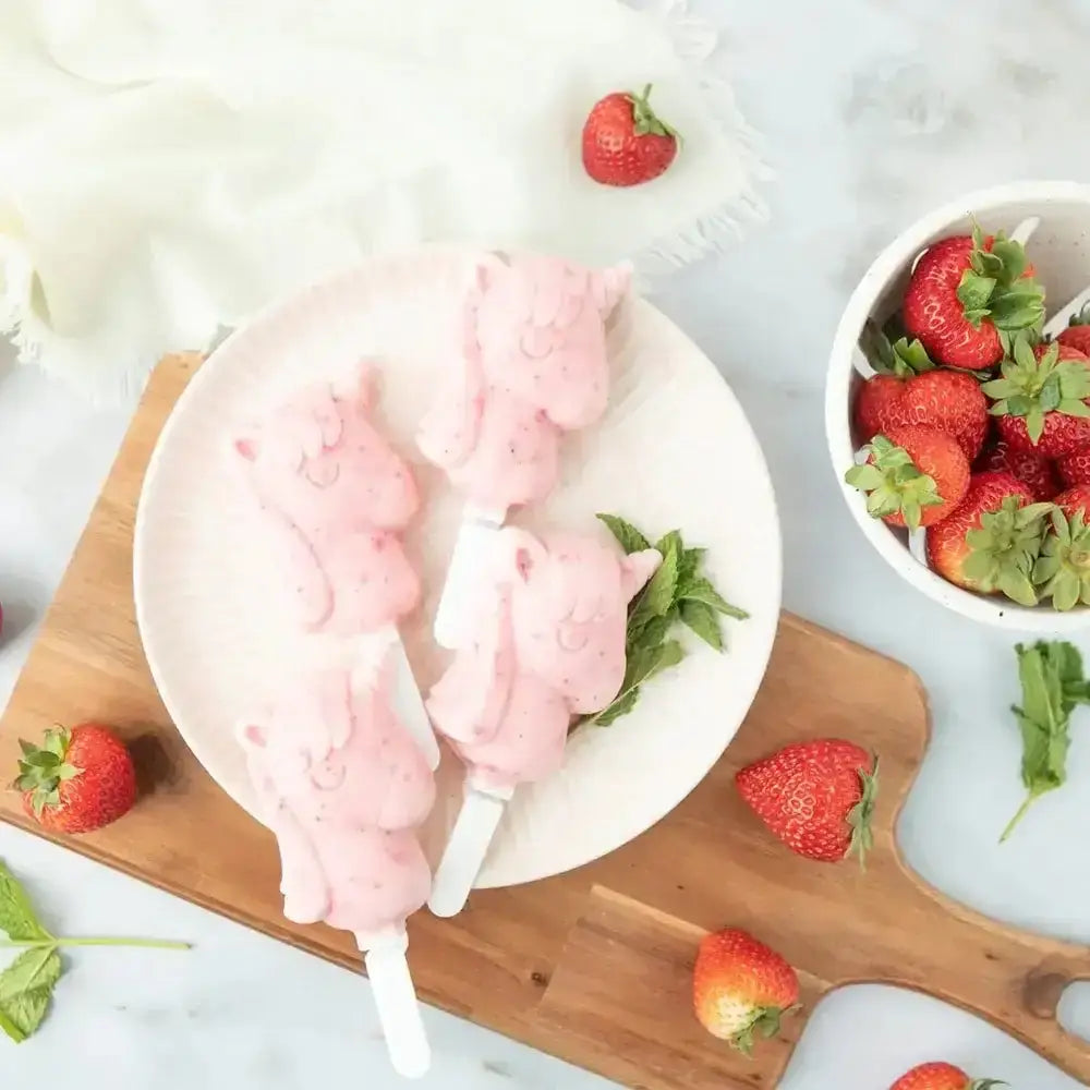 Pink ice cream lollipops on a white plate with strawberries on a wooden board.