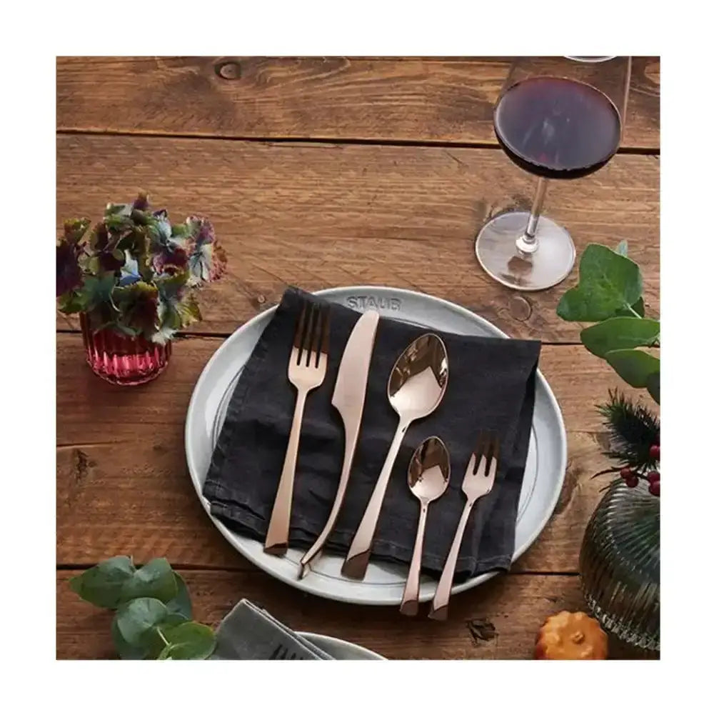 Rose gold cutlery set on a white plate with a dark napkin, placed on a wooden table with a glass of red wine.