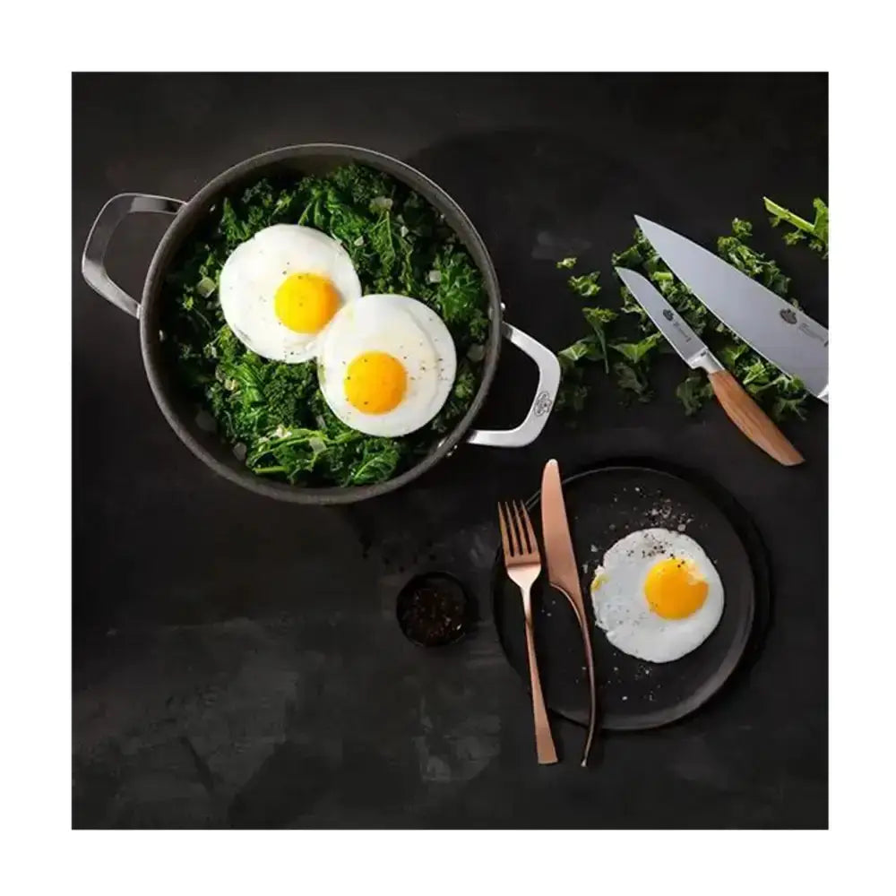 Fried eggs with greens in a pan, served with a knife and fork on a dark surface.