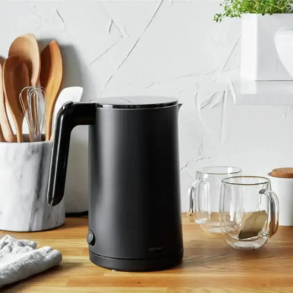 Black electric kettle on a kitchen counter with wooden spoons and glasses in the background.