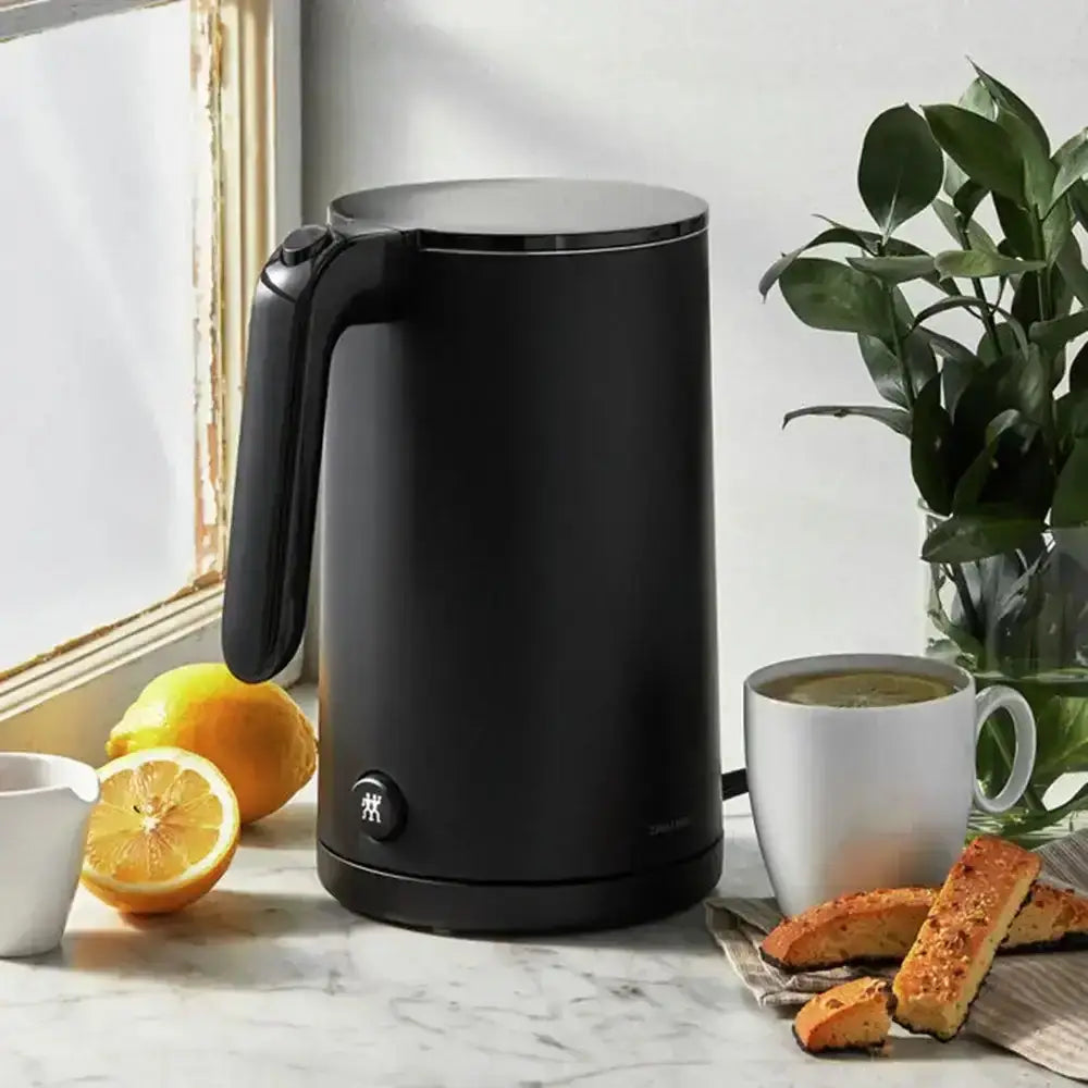 Black electric kettle on a marble countertop with a cup of coffee, biscuits, and lemons.